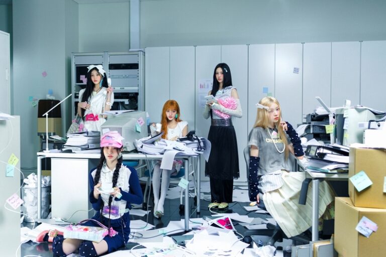 Five K-pop idols posing in a chaotic office setting with scattered papers, boxes, and vintage tech equipment.