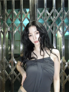 Karina with long, wavy black hair wearing a dark gray off-shoulder top stands in front of a metallic crisscross-patterned gate, with teal lighting in the background.