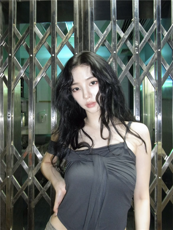 Karina with long, wavy black hair wearing a dark gray off-shoulder top stands in front of a metallic crisscross-patterned gate, with teal lighting in the background.