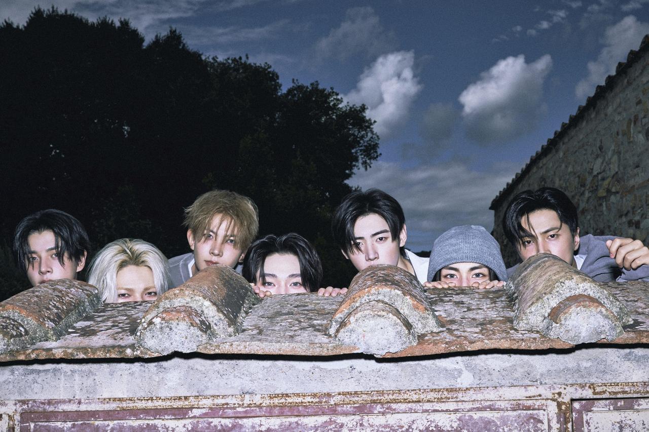 ENHYPEN members peeking over a rustic stone wall at twilight with dramatic sky and vintage ambiance.