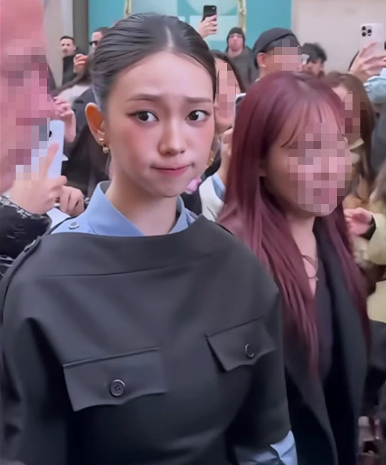 Karina with tied-back hair wearing a black outfit over a light blue collared shirt stands in front of turquoise doors, surrounded by a crowd holding up smartphones.