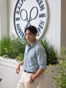 SEVENTEEN’s DK poses in a light blue button‑up shirt and white pants in front of a Ralph Lauren Australian Open sign featuring crossed tennis rackets. He stands confidently with one hand in his pocket and the other resting on a panel, surrounded by greenery in a modern setting.