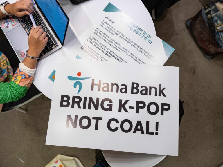 A protest sign on a table reads “Hana Bank BRING K-POP NOT COAL!” next to a laptop, symbolizing Kpop fans’ climate campaign against coal financing.