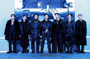 Seven BTS members stand in front of a bullet-riddled vault door glowing blue, dressed in coordinated black outfits with leather, fur, and chains, surrounded by scattered gold bullet casings.