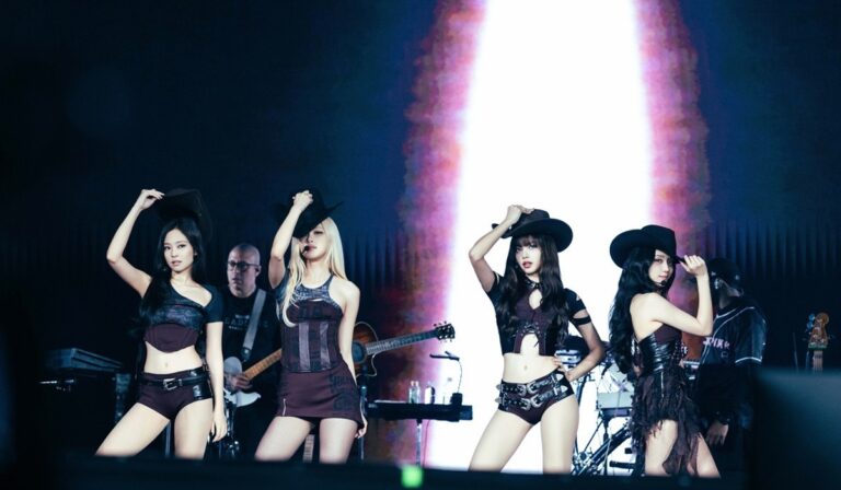 BLACKPINK live performance with four members in dark outfits and cowboy hats, striking synchronized poses on stage with band and illuminated backdrop.”