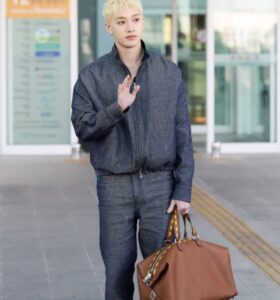 Bang Chan of Stray Kids at Incheon International Airport en route to London Fashion Week. He is wearing a dark denim jacket and jeans set, with short bleached blonde hair, holding a large brown Fendi leather bag with yellow and brown striped handles. He waves to fans while standing in front of the terminal entrance.