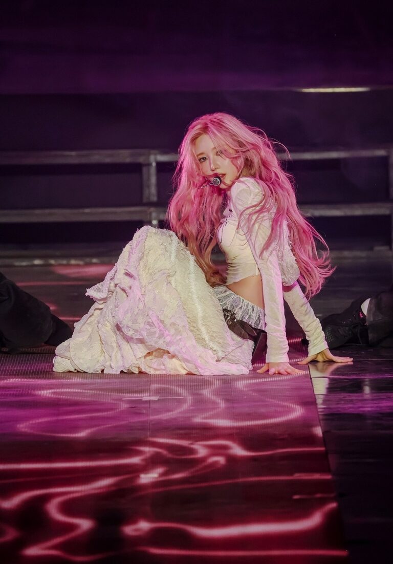 Gaeul with pink hair sings on stage in white outfit under pink and purple lights.