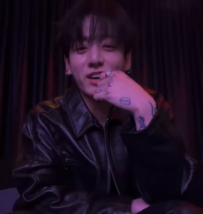 Jungkook sits against dark curtains in a black leather outfit, hands interlaced, under moody red‑purple lighting during a Weverse live.
