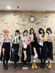 IVE members posing with flower bouquets in front of a brick wall and clock, celebrating a win backstage.