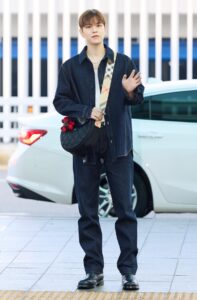 Stray Kids’ Seungmin at Incheon International Airport en route to London Fashion Week. He is dressed in a Burberry-inspired outfit with a dark denim shirt and jeans, accessorized with a black quilted crossbody bag featuring a beige plaid strap and red plush charms.