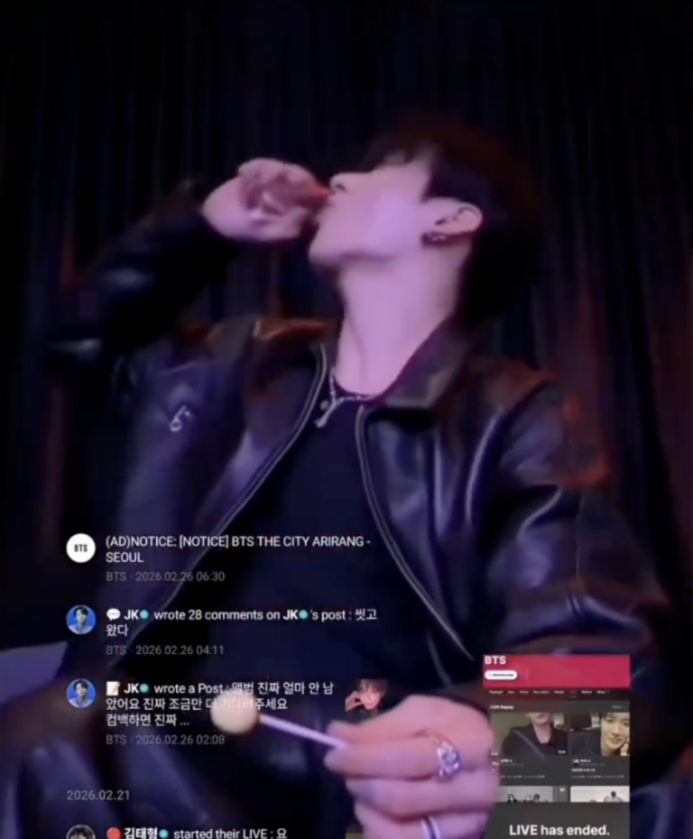 A BTS member Jungkook sits in front of a dark curtain backdrop during a Weverse live, wearing a black leather jacket and holding a lollipop.