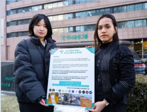 Lee Dayeon (left) and an Indonesian activist stand in front of Hana Financial Group headquarters in downtown Seoul, holding a large placard urging Hana Bank to stop financing palm oil companies accused of deforestation and human rights violations, highlighting global Kpop fans’ demand for sustainable finance.