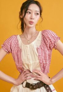 Giselle stands against an orange background wearing a cream dress layered with a red-and-white checkered cropped top.