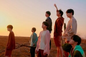 BTS members pose in a serene field at sunset, dressed in earthy pastel tones.