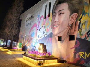 A large mural of BTS leader RM is displayed on a building wall in Goyang, illuminated at night with colorful street art elements.