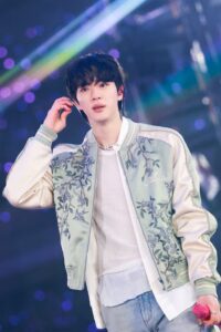 BTS Jin performs on stage in a floral embroidered jacket, holding an in-ear monitor amid colorful lights.