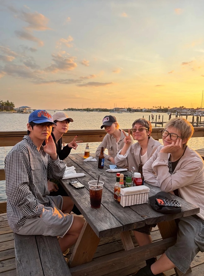 BTS members sit together at a waterfront table in Tampa during sunset.