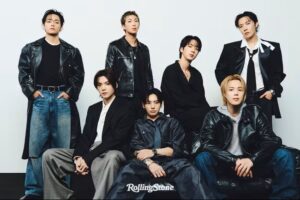 BTS members pose together in a studio wearing coordinated dark outfits, seated and standing against a white background.