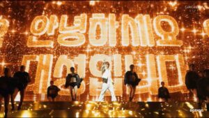 BIGBANG performs on a brightly lit Coachella stage with a golden LED backdrop displaying Korean text.