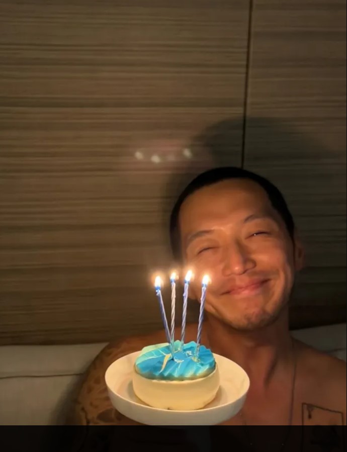 Byun Yo‑han sits indoors holding a dessert with blue icing and four lit candles.