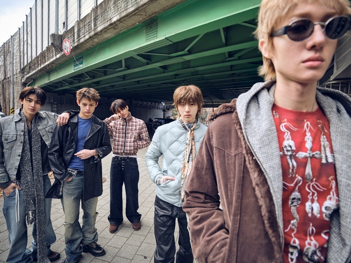 CORTIS members pose in layered street‑style outfits under a green overpass in an urban setting.
