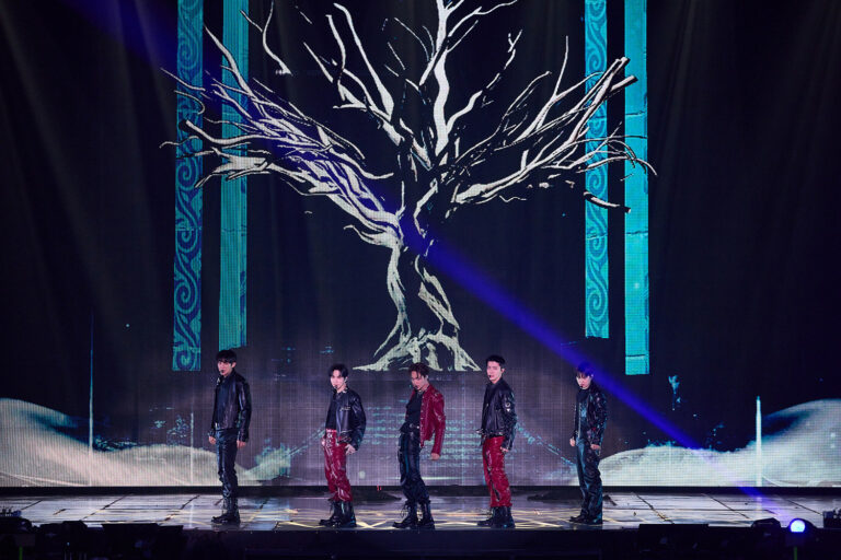 Five EXO members perform on stage in Seoul, wearing black and red leather outfits, with a digital backdrop of a tree illuminated by blue and purple lights.