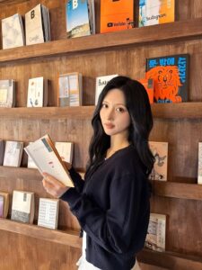 Gaeul stands in front of a wooden bookshelf filled with books and magazines, holding an open book.