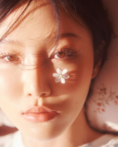 Hwasa’s face in close‑up with a small white flower placed on her cheek under soft lighting.