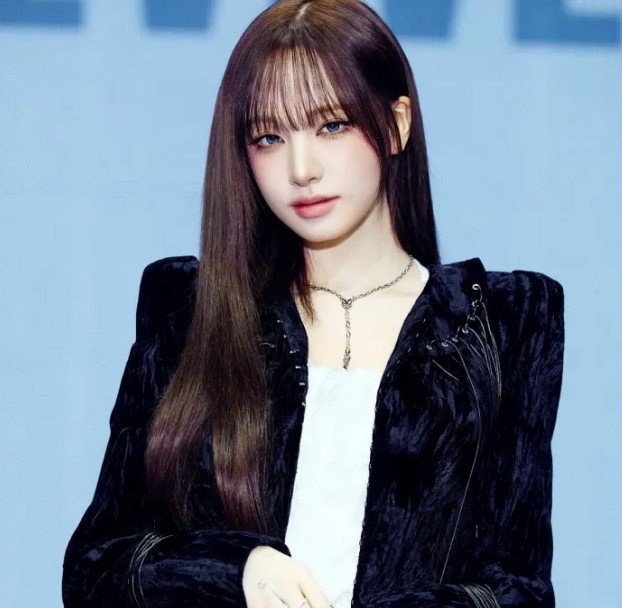 Liz stands against a light blue background wearing a black textured jacket and silver chain necklace.