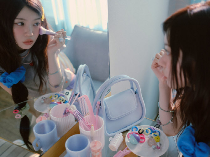 ILLIT’s Moka sits at a vanity table applying makeup, surrounded by accessories and soft lighting.