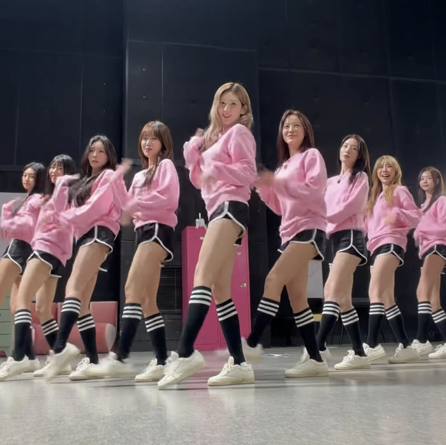I.O.I members rehearse a synchronized dance routine in matching pink sweatshirts and black shorts.