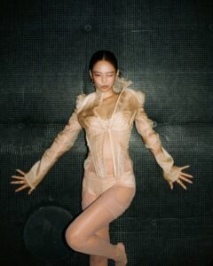 Jennie of BLACKPINK lies on a dark surface wearing a sheer, light-colored layered outfit with extended arms.