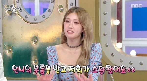Jeon Somi speaks on a Korean TV talk show, wearing a light blue floral dress with puffed sleeves.