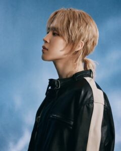 Profile portrait of BTS member Jimin with blond layered hair tied in a short ponytail, wearing a black leather jacket with beige stripe detail against a soft blue background.