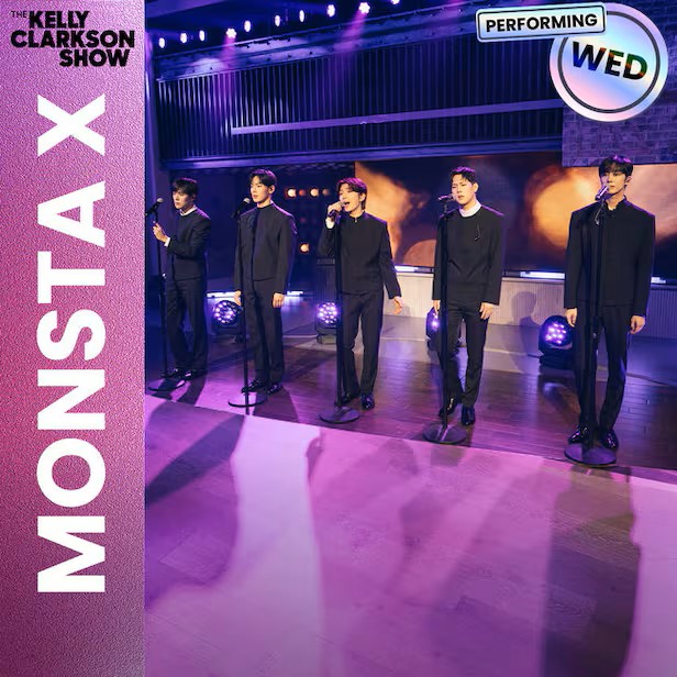 MONSTA X members perform on stage in coordinated dark outfits, standing at microphone stands under purple and amber lighting.