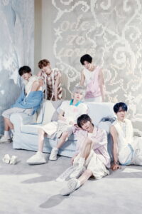 Six NCT WISH members pose together in a pastel-themed room, dressed in soft-toned outfits around a light blue couch.