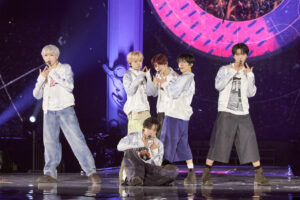 NCT WISH members perform in coordinated light jackets before a vibrant circular stage display.