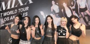 NMIXX members pose in black and gray outfits before a backdrop promoting their Los Angeles world tour stop.