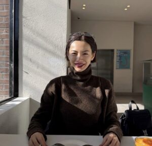 Nana sits at a table near a window, wearing a dark turtleneck with sunglasses on her head.