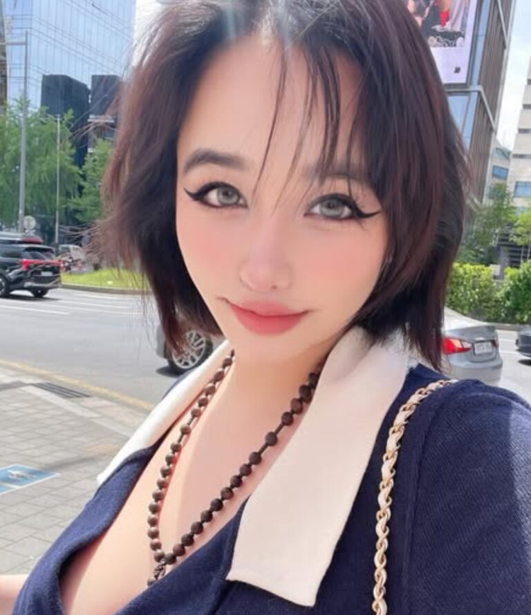 Park Bom takes a bright selfie indoors, wearing a bold navy V‑neck top with a beaded necklace.