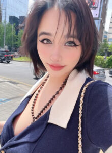 Park Bom takes a bright selfie indoors, wearing a bold navy V‑neck top with a beaded necklace.