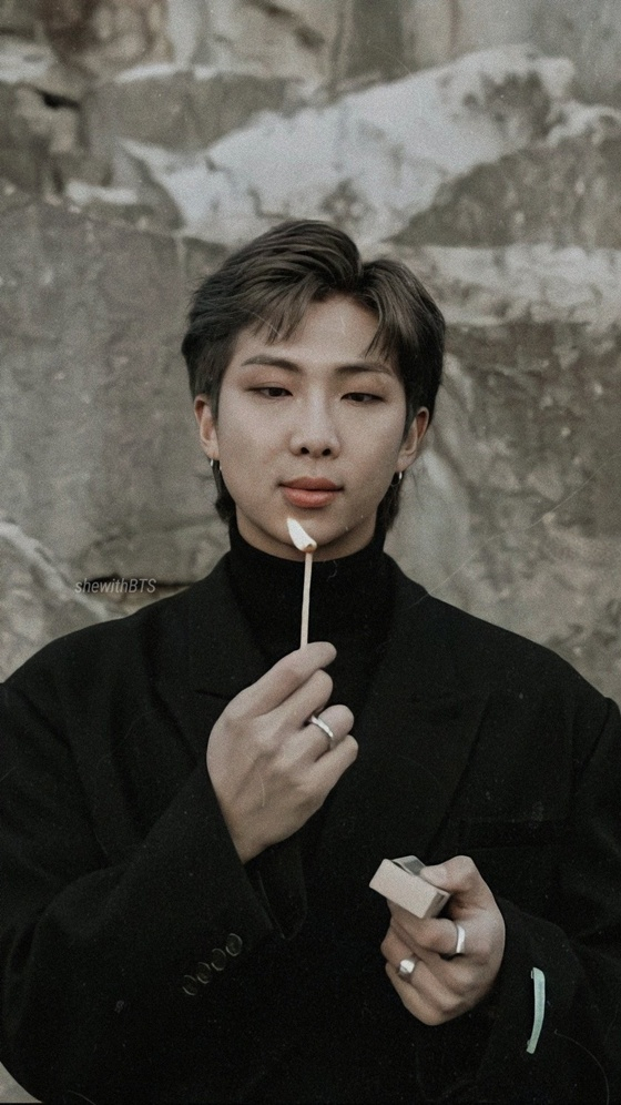 RM stands outdoors in front of a rocky background, wearing a black turtleneck and blazer, holding a lit matchstick and matchbox.