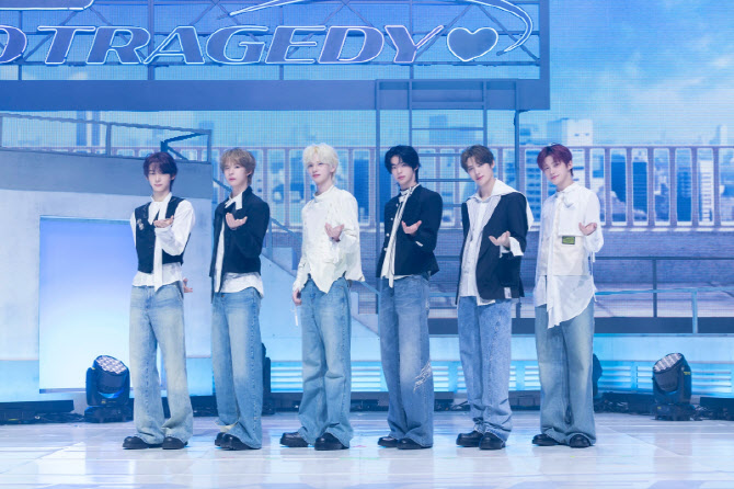 TWS members perform on stage in coordinated outfits under a glowing sign reading ‘TRAGEDY♥.’”