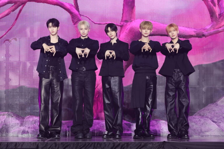 TXT’s five members stand on stage in black outfits against a vivid pink and purple backdrop with stylized tree branches, posing with synchronized hand gestures.