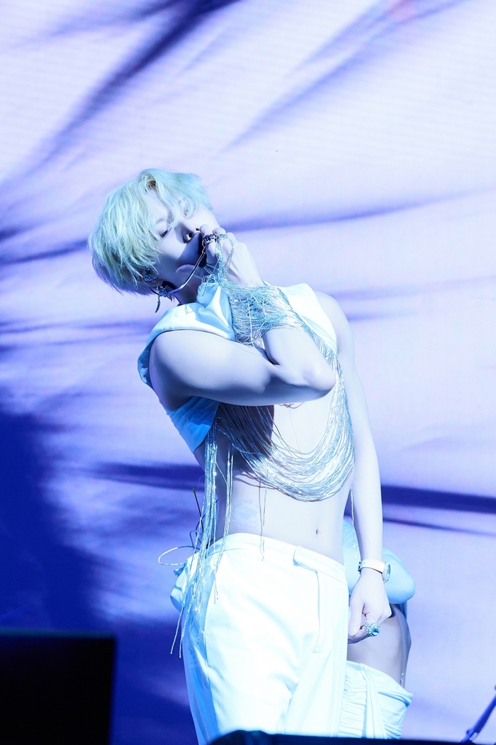 Taemin poses on stage in a white outfit with metallic fringe, illuminated by blue and purple lighting.
