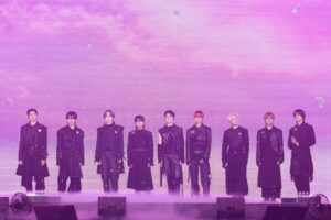 &TEAM perform on stage in coordinated black outfits against a pink and purple lit backdrop.