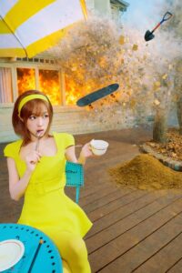 YENA sits calmly at a turquoise table in a bright yellow dress, sipping tea while a house explodes in the background.