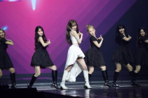 YENA performs on stage in a white outfit, surrounded by dancers in black lace dresses against a pink-lit backdrop.