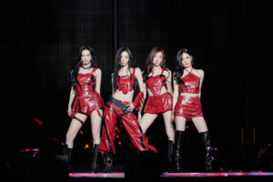 aespa members stand confidently in formation wearing shiny red outfits and black boots under bright stage lights.