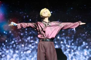 Taemin performs on stage in a shiny maroon outfit, arms outstretched under glittering lights.